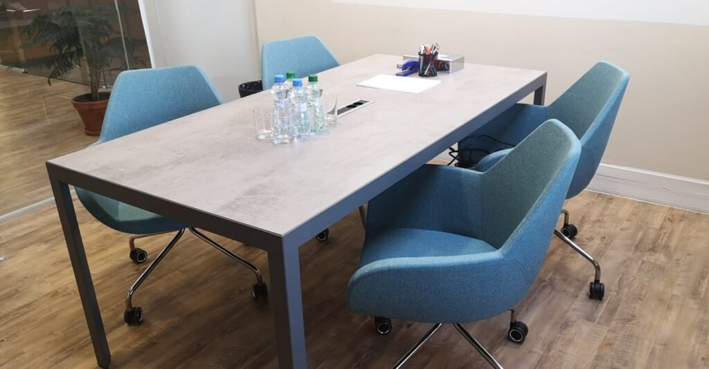 Návrh kancelářských prostor pro společnost 24 zahrnuje konferenční stůl a pohodlné modré židle na kolečkách pro pracovní schůzky.